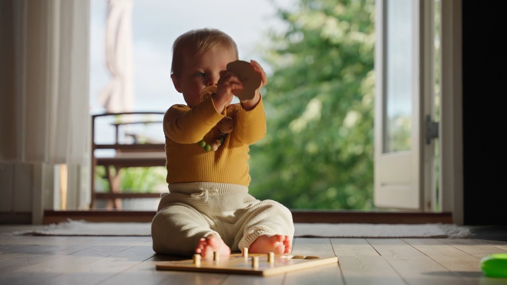 Beba sedi na podu i drži drvenu igračku u rukama, ispred nje se nalazi slagalica