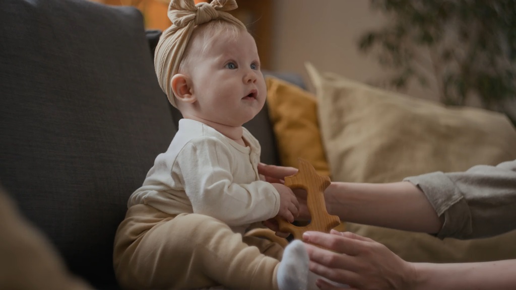 Beba sedi na sofi uz podršku roditelja, u rukama drži drvenu igračku